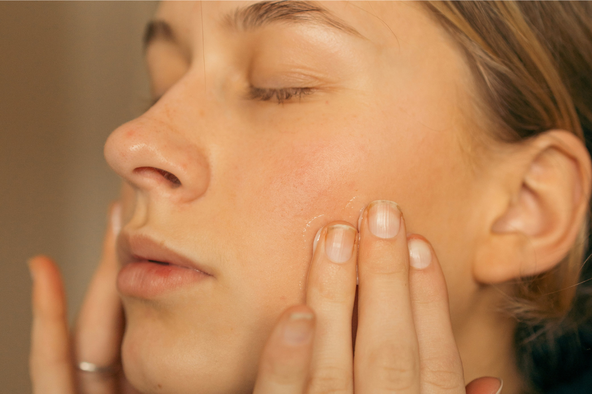 Close-up of person gently applying skincare product to cheek with fingertips. Natural lighting, eyes closed, relaxed expression, minimal background. Focus on skin texture and hands.