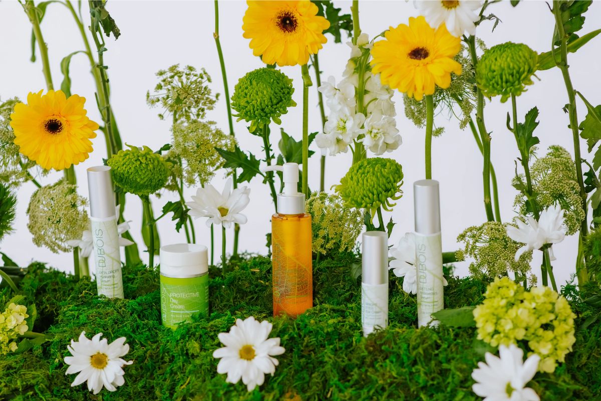 Dubois skincare products displayed on green moss with yellow and white flowers, including a cream jar, spray bottle, and serum tubes in a bright, botanical setting.