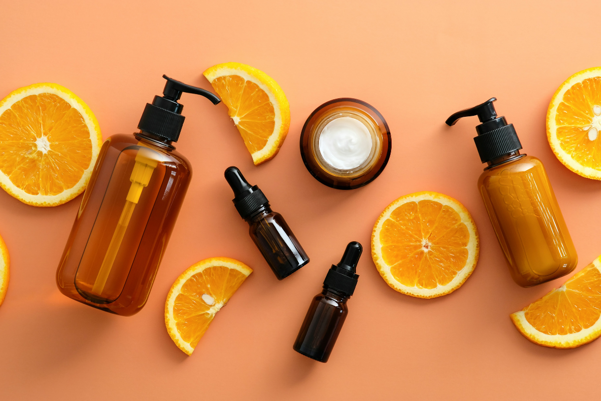 Assorted amber bottles and dropper jars with sliced oranges on a peach background. Includes pump dispensers and cream jar, suggesting skincare or cosmetic theme. Bright, fresh composition.