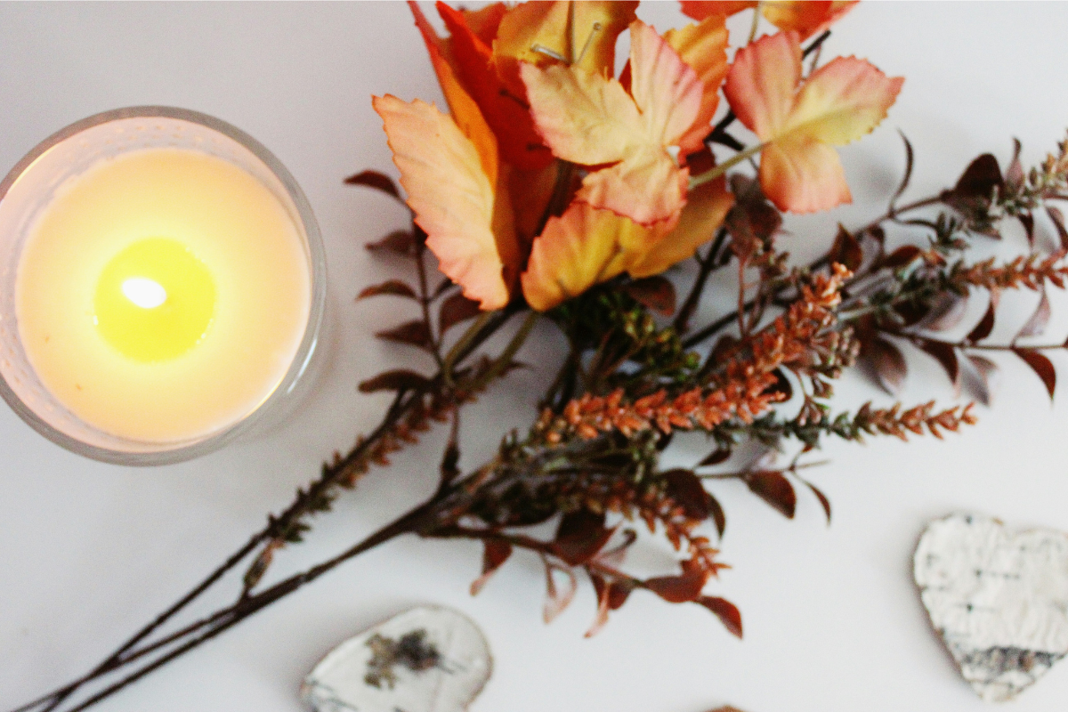 Top view of a lit candle next to autumn leaves and dried branches on a white surface. Two pieces of bark are visible in the corner.