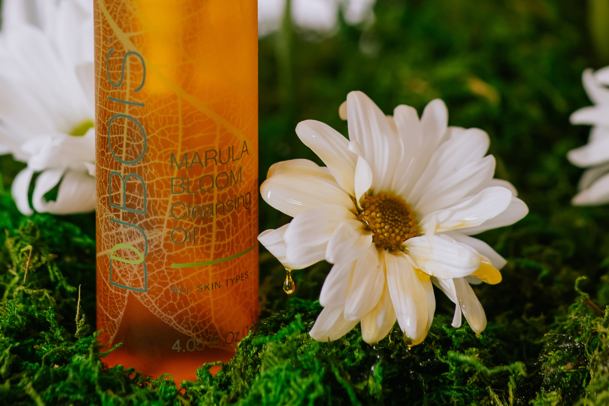 Bottle of Marula Bloom Cleansing Oil on lush green moss, complemented by white daisies. Natural, fresh setting, highlighting the product's connection to nature. Suitable for all skin types.