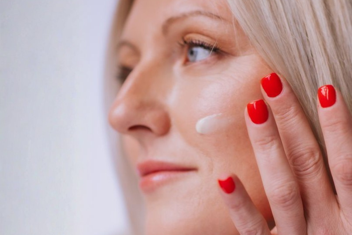 Close-up of a woman applying skincare cream to her cheek with her fingertips. She has red nails, light hair, and is smiling slightly. Natural lighting, showcasing skincare routine.