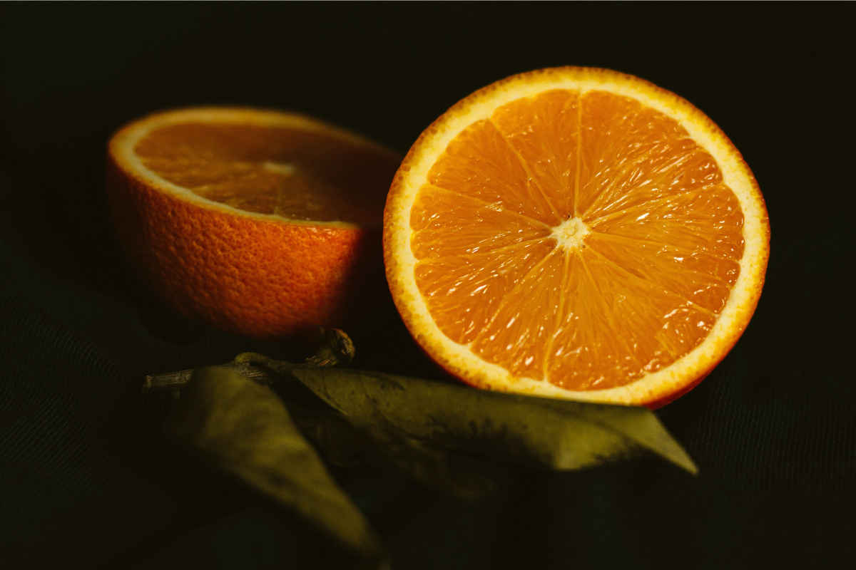 Close-up of two orange halves resting on a dark surface. The vibrant orange color contrasts with the deep background, highlighting the juicy texture and freshness of the fruit.