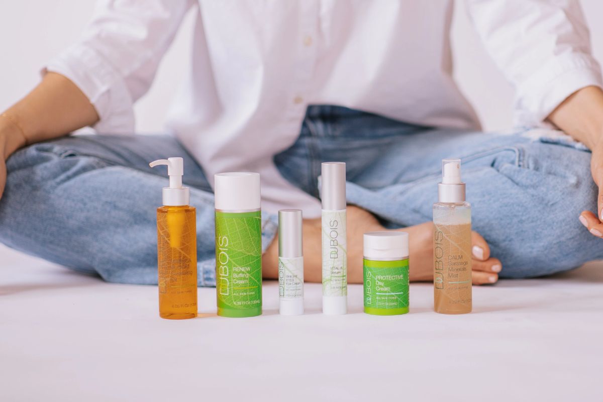 Person sitting cross-legged in jeans and white shirt behind a lineup of Dubois skincare products, including pump bottles, spray mist, creams, and serums on a light background.