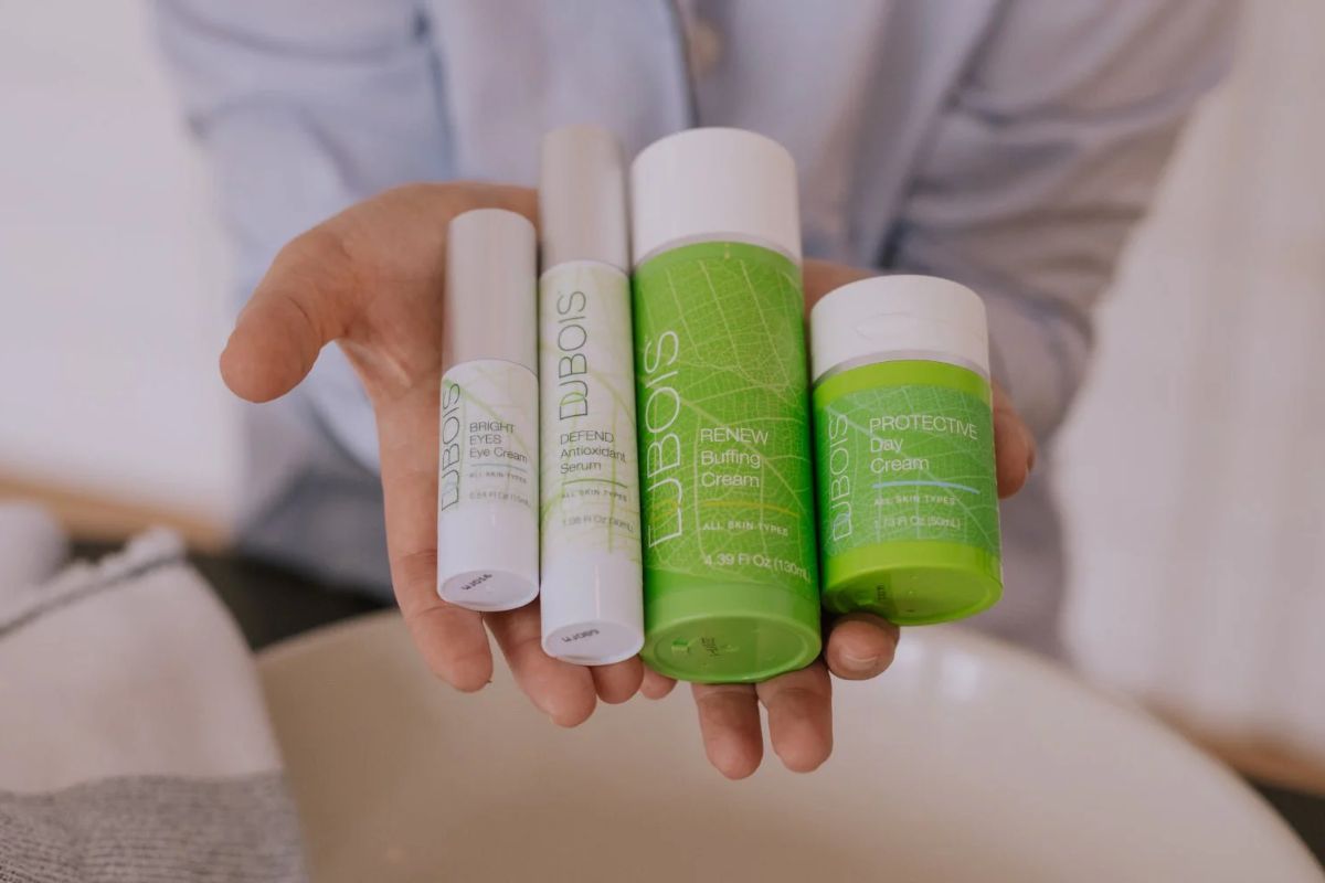 Hand holding four Dubois skincare items: Bright Eyes Eye Cream, Defend Antioxidant Serum, Renew Buffing Cream, and Protective Day Cream, with green labels and white caps.