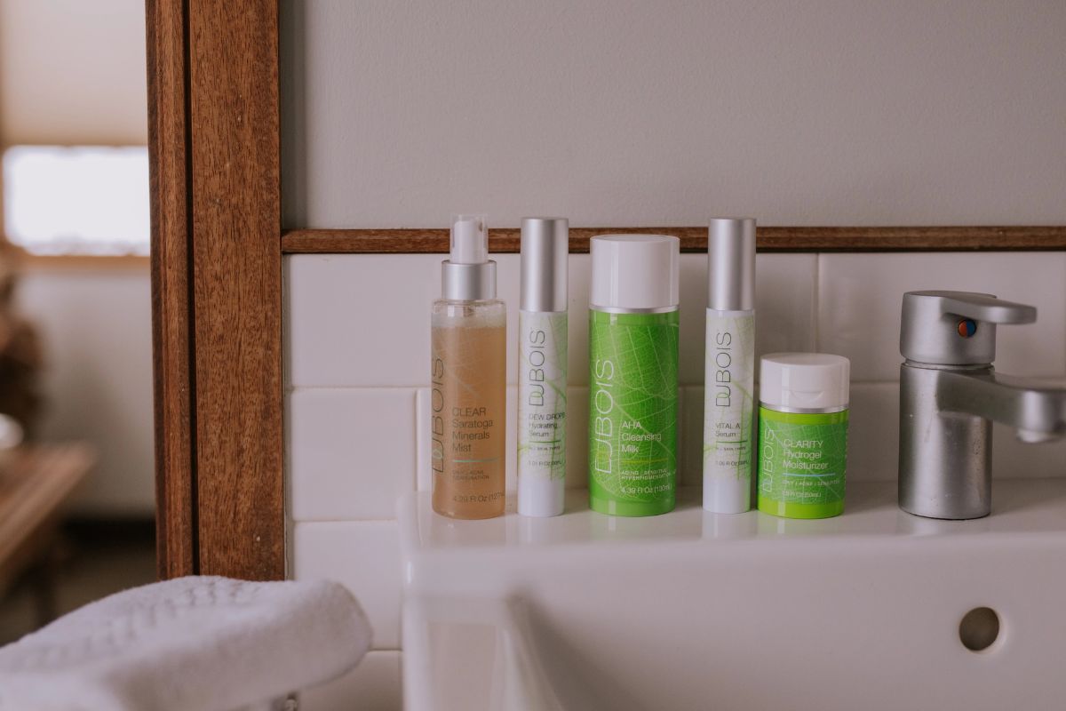 Dubois skincare products lined up on a bathroom sink beside a faucet and mirror, including Saratoga Minerals Mist, Dew Drops Serum, AHA Cleansing Milk, Vital A Serum, and Clarity Hydrogel Moisturizer.