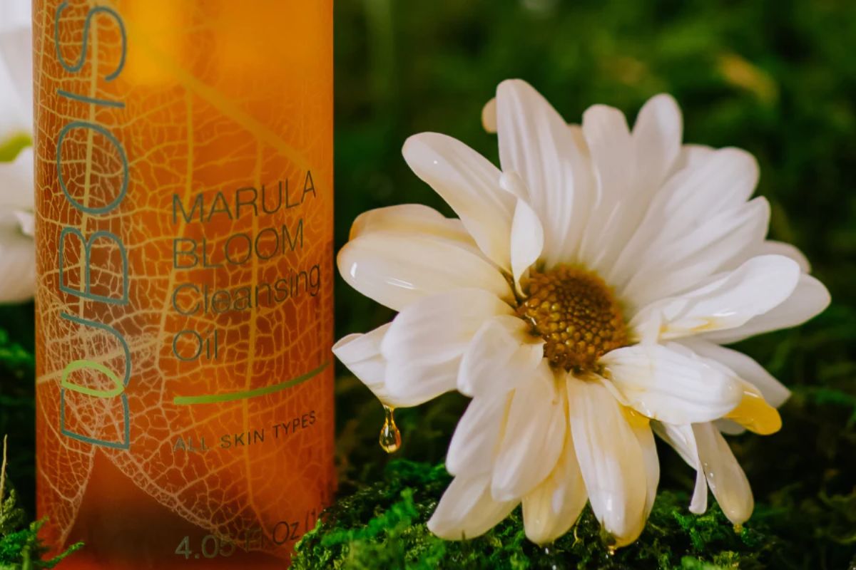 Bottle of Marula Bloom Cleansing Oil on lush green moss, complemented by white daisies. Natural, fresh setting, highlighting the product's connection to nature. Suitable for all skin types.