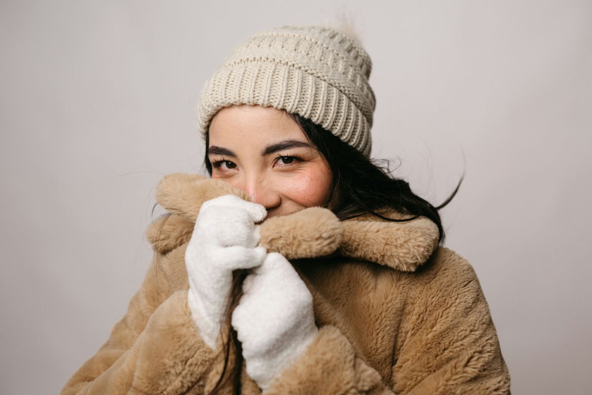 Person in a beige winter coat and knit beanie holding a white scarf over their face, against a plain light background.