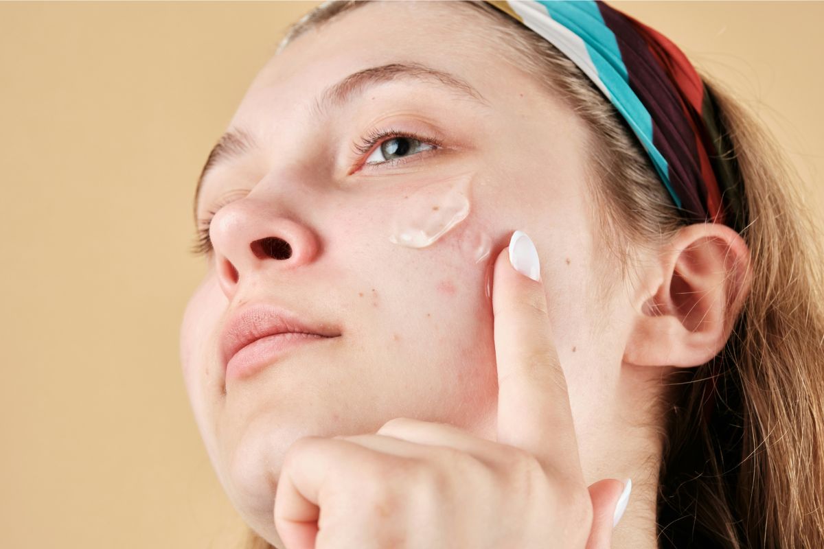Close-up of person with headband applying a small dab of face cream to cheek with fingertip, against a warm beige background.