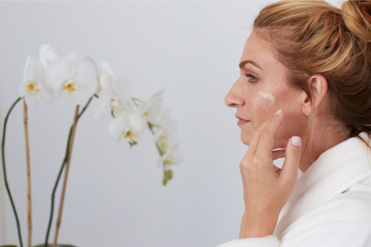 Person in a white robe with hair in a bun touches their chin, with white orchids in the background. Soft, minimal spa-like setting.