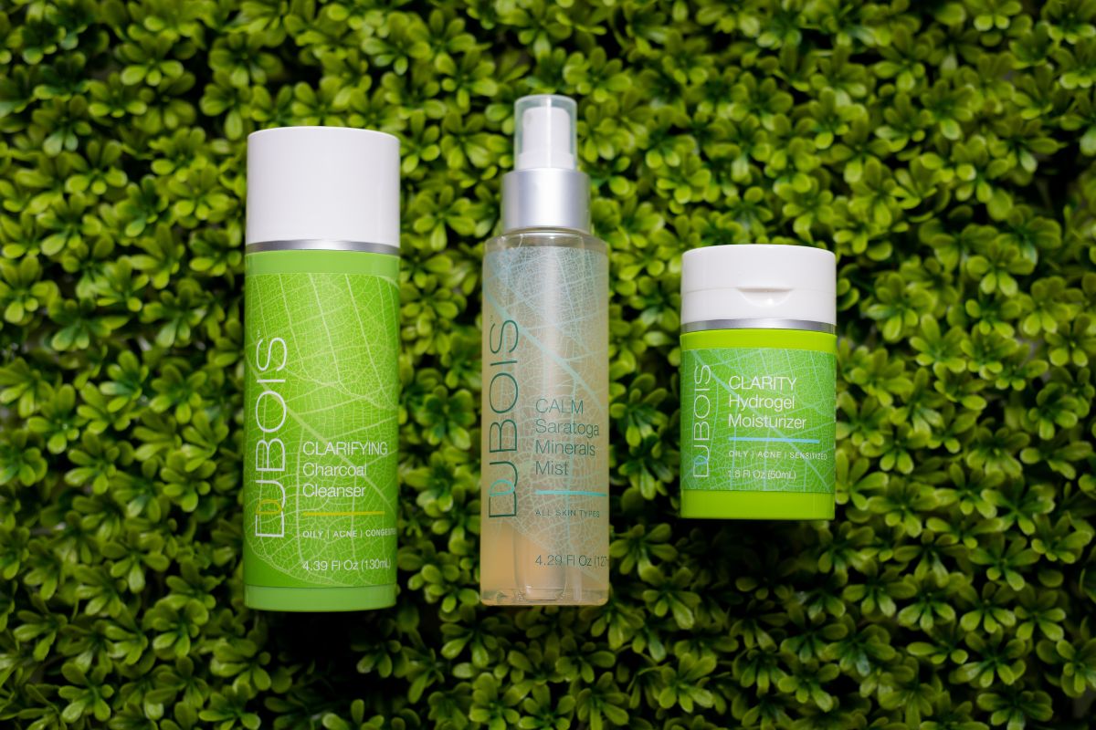 Three Dubois skincare products on a leafy green background: Clarifying Charcoal Cleanser, Calm Saratoga Minerals Mist, and Clarity Hydrogel Moisturizer.