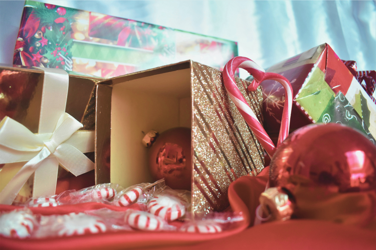 Festive scene with open gift boxes, candy canes, peppermint candies, and red ornaments on a red fabric. Glittery box and holiday-themed wrapping in background. Holiday decorations.