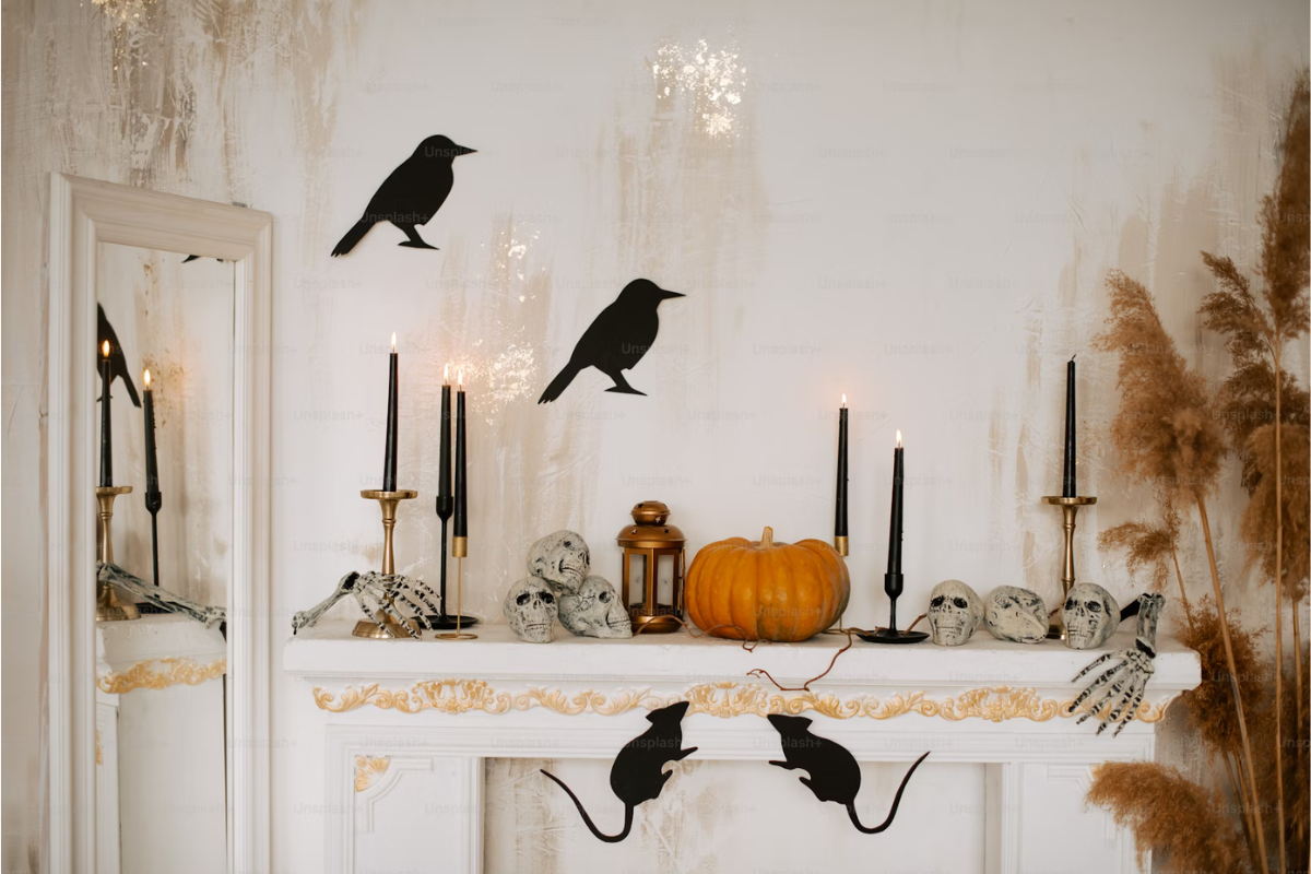 Decorative Halloween mantel with black candles, skulls, pumpkin, crow silhouettes, and black rat cutouts. Mirror reflects elements; dried plants on side.