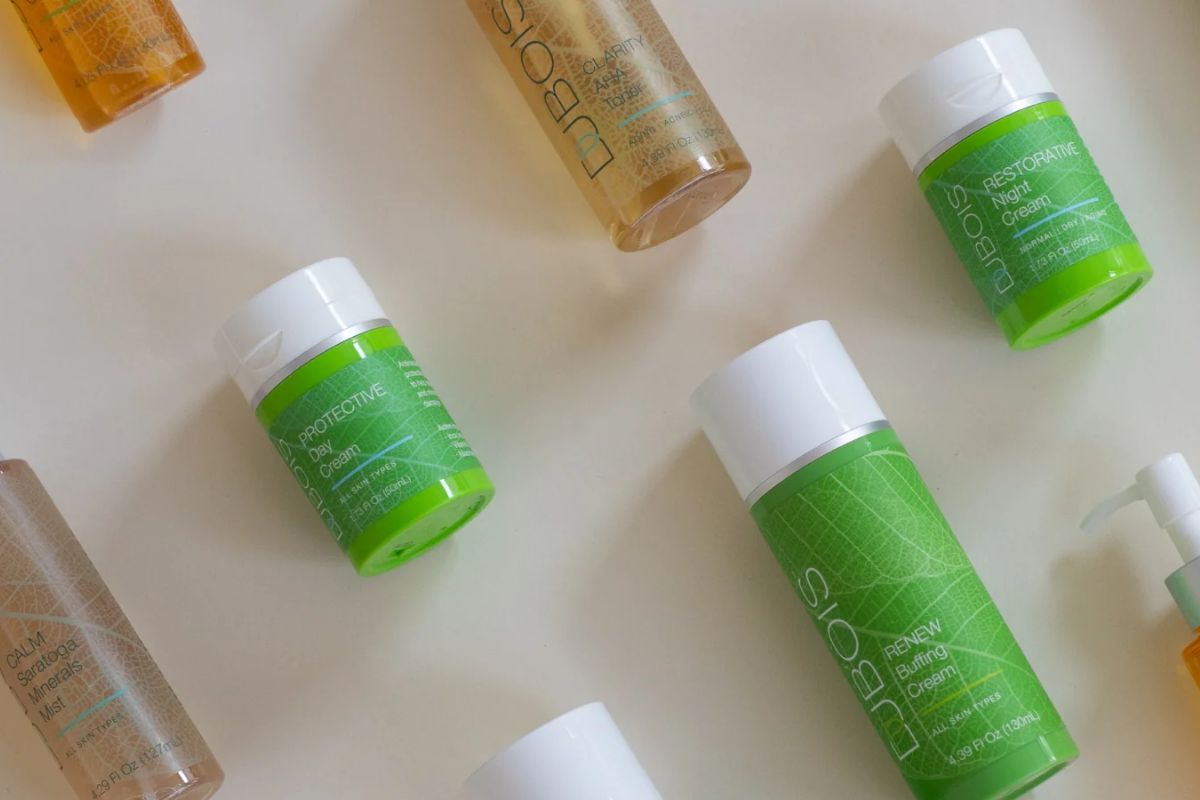 Flat lay of Dubois skincare products on a light surface, including Renew Buffing Cream, Protective Day Cream, Restorative Night Cream, Calm Saratoga Minerals Mist, and Clarity AHA Cleansing Milk.