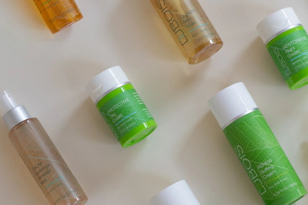 Flat lay of Dubois skincare bottles and jars on a light background, including Calm Saratoga Minerals Mist, Clarity AHA Cleansing Milk, Renew Buffing Cream, Protective Day Cream, and Restorative Night Cream.