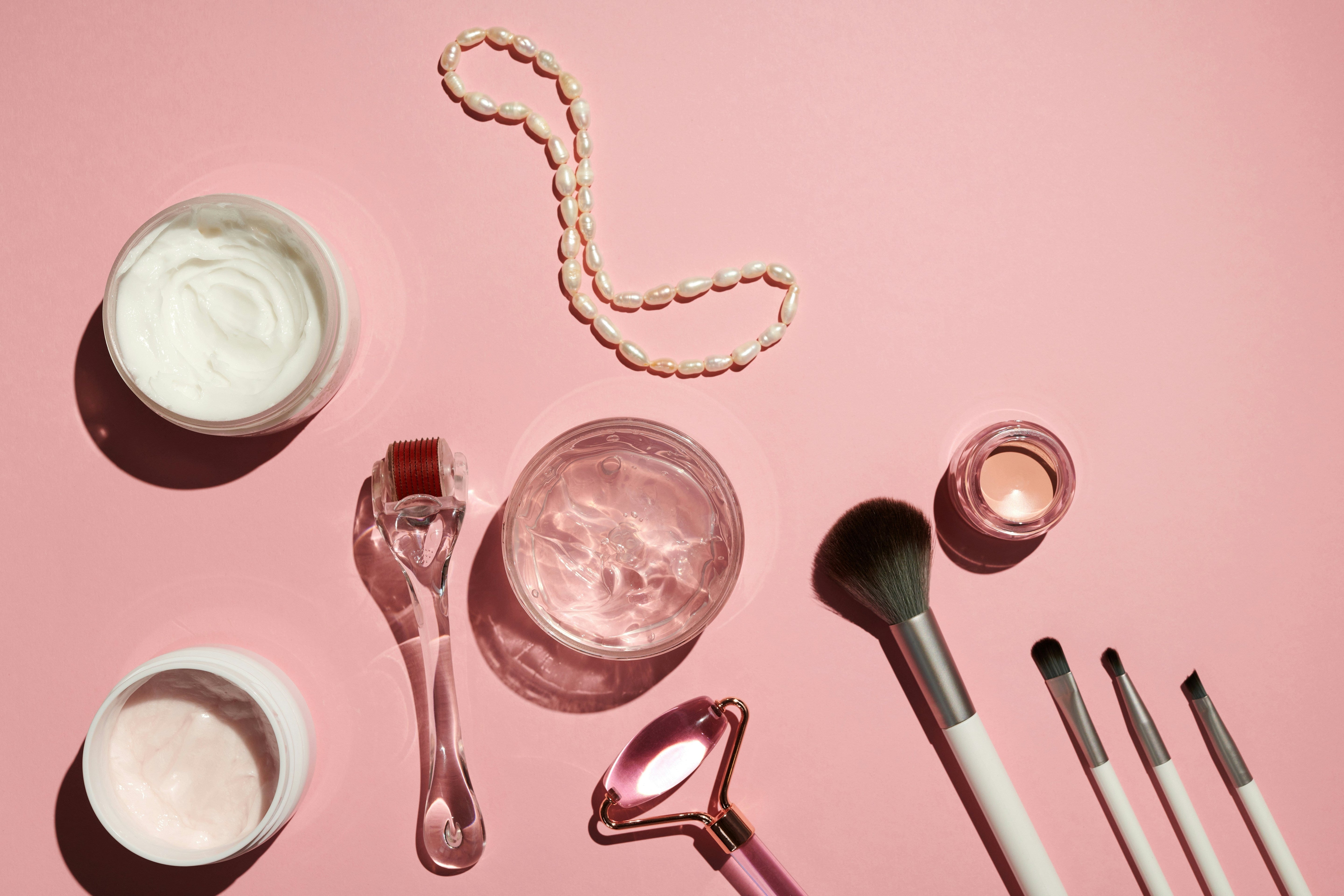 Flat lay of skincare and makeup items on pink background, including cream jars, a jade roller, derma roller, makeup brushes, small round mirror, and a string of pearls. Stylish composition with soft shadows.